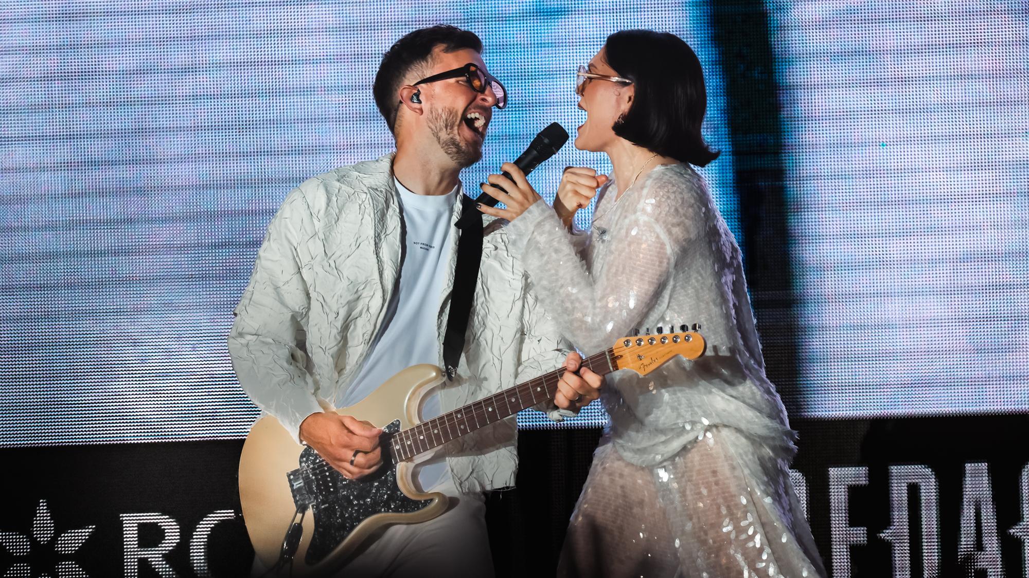 To sangere synger sammen på scenen, én spiller elguitar