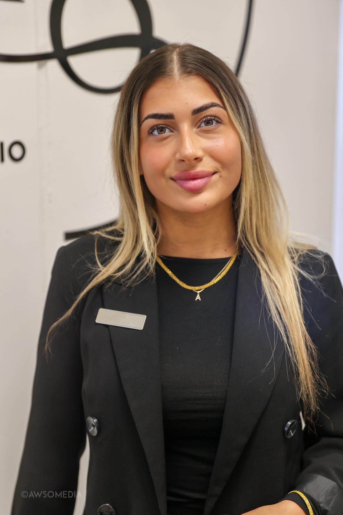 Kvinde med blondt hår i sort blazer smiler foran logo-væg